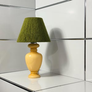 May include: A table lamp featuring a smooth, glossy, pale yellow ceramic base with a rounded, vase-like silhouette. The lamp is topped with a conical lampshade wrapped in textured, dark olive green yarn, creating a distinct contrast in color and material.