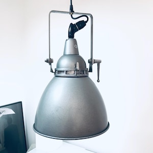 Puede incluir: Lámpara colgante industrial plateada con pantalla en forma de cúpula. La lámpara tiene un marco de metal y un cable negro. El diseño recuerda la iluminación de fábrica vintage.