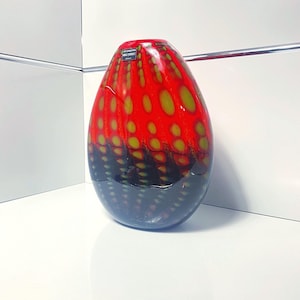 May include: A decorative glass vase with a rounded shape. The top half is red with yellow-green spots, transitioning to a black base. The vase has a glossy finish and a label near the top.