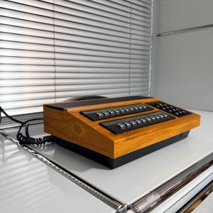 May include: A retro wooden telephone console with black buttons and a black base. The console features a vintage design with multiple rows of buttons and a sleek, angled shape. A black cord extends from the side.