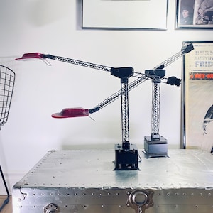 Peut inclure: Deux lampes de bureau noires avec des panneaux lumineux rouges. Les lampes ont un design unique avec un bras en treillis et une base carrée. Elles sont posées sur un coffre en métal argenté, avec des œuvres encadrées en arrière-plan.