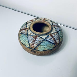 May include: A small, round ceramic vase with a wide opening. The vase features a geometric pattern in shades of blue, brown, and white. The rim and base are a darker brown, and the interior is a lighter tan color. The vase is likely handmade.