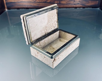 Op de afbeelding: Een klein, rechthoekig doosje van beige travertijn met een zilveren metalen rand. Het doosje is open en laat een lege binnenkant zien.