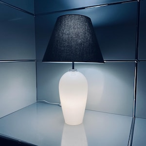 Può includere: Una lampada da tavolo in vetro smerigliato bianco con paralume nero. La lampada ha una base arrotondata e un paralume in tessuto grigio scuro. La lampada è collegata a una presa a muro. La lampada è su una superficie bianca.