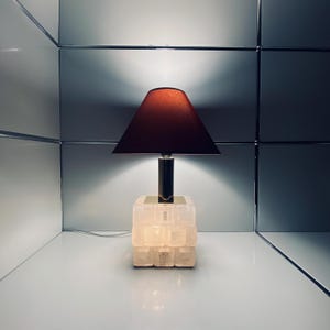 May include: A contemporary table lamp featuring a burgundy lampshade and a textured, light-coloured base. The lamp has a dark metal stem and a square base. The lamp is plugged into an electrical outlet.