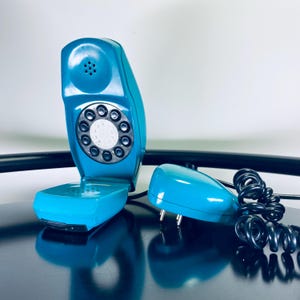 May include: A teal blue rotary phone with a separate handset. The phone has a round dial with black numbers and a speaker on the top. The handset is also teal blue and has a cord.