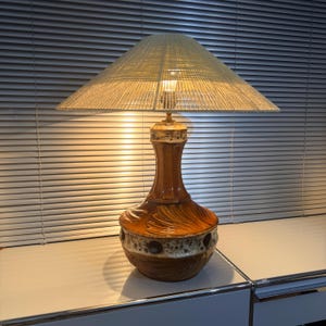 Puede incluir: Lámpara de mesa vintage con base de cerámica marrón y pantalla beige tejida. La base tiene forma bulbosa con diseño texturizado y banda de agujeros oscuros. La pantalla es cónica y proyecta un brillo cálido. Mide unos 60 cm de alto.