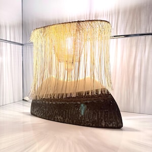 Könnte beinhalten: Eine einzigartige Tischlampe mit einem strukturierten, dunkelbraunen Sockel und einem gewebten, gefransten Schirm. Der Schirm besteht aus einem hellen Material, und die Lampe strahlt ein warmes, einladendes Licht aus. Das Design ist modern und künstlerisch.