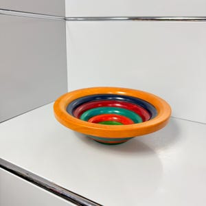 Puede incluir: Un cuenco circular de madera con un diseño anidado y multicolor. El cuenco muestra anillos concéntricos de color naranja, azul oscuro, rojo y verde, con un acabado liso y pulido que resalta la veta natural de la madera en toda la estructura.