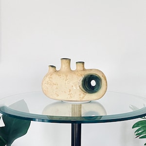 Può includere: Un vaso in ceramica con un designo astratto unico. Il vaso è di colore crema con accenti verdi e blu e ha tre aperture per i fiori. È posto su un tavolo di vetro.