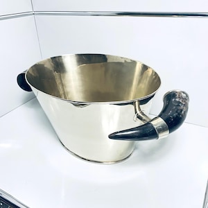 Puede incluir: Una cubitera plateada con forma ovalada ensanchada. Cuenta con un asa de cuerno negro con detalles plateados en un lado y un pequeño asa negra en el otro. El interior es reflectante.