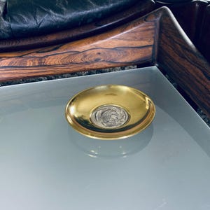 May include: A small, round, gold-colored dish with a silver-colored design of a woman's head in profile. The dish is on a glass surface.