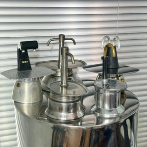 Puede incluir: Una colección de cuatro cafeteras italianas de metal. Una presenta un mango negro con el texto "luxa" en el lateral y una placa de metal martillado. Las otras son de tono plateado con varios diseños de boquilla, incluyendo tubos de metal curvos.