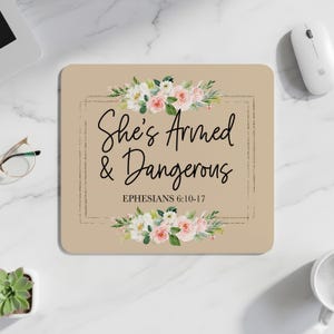 Peut inclure: Un tapis de souris beige avec l'inscription "She's Armed & Dangerous" en écriture noire, encadrée d'une ligne pointillée. Des compositions florales avec des roses roses et blanches et de la verdure ornent le haut et le bas. Le verset biblique Éphésiens 6:10-17 est en dessous.