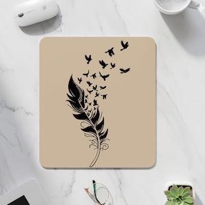 May include: A beige square mouse mat with a black feather design and birds in flight. The feather has detailed curves and swirls. The birds are shown in silhouette, suggesting movement and freedom. A decorative item.