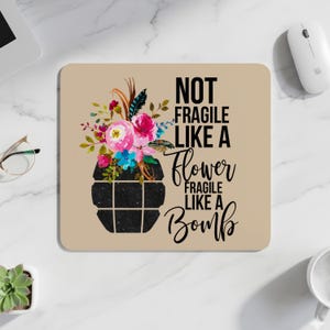 May include: A beige mousepad features a floral design with pink, blue, and red flowers emerging from a black, grid-patterned shape. The text reads "NOT FRAGILE LIKE A FLOWER FRAGILE LIKE A BOMB."