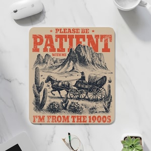 May include: A beige mousepad with a vintage western design. The image features a horse-drawn carriage, cacti, and a mountain range. Text reads "PLEASE BE PATIENT WITH ME I'M FROM THE 1900S" in red and black.