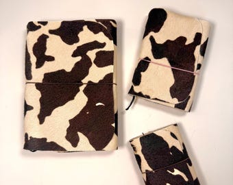 May include: Three rectangular journals with a cow print pattern featuring dark brown and cream patches. Each journal is secured with a thin elastic band wrapped horizontally around the center, holding the covers closed in a simple, functional design.