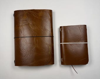 May include: Two brown leather journals with a crinkled texture. The larger journal features a black elastic band closure, while the smaller journal is secured with a white elastic band. Both items have rounded corners and a simple, functional design.