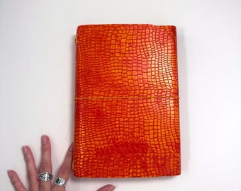 May include: A rectangular journal with a vibrant orange and red crocodile-embossed texture. The cover features a glossy finish and is secured by a thin, horizontal orange elastic band that wraps around the center of the notebook.
