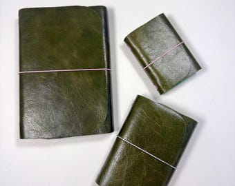 May include: Three olive green leather journals with a textured finish. The largest journal is secured with a pink elastic band, while the two smaller journals are held closed by thin pink and white elastic bands respectively.