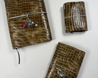 May include: Three journals with a brown crocodile-embossed texture. The largest and medium journals feature silver elastic bands with dangling charms, including a silver dragonfly and colorful beads. The smallest journal has a plain silver elastic band.
