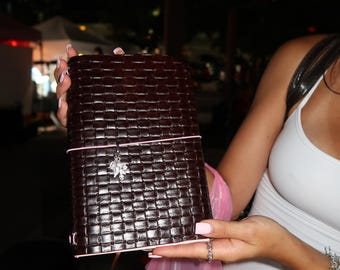 May include: A dark brown rectangular notebook with a woven leather-like texture. The cover is secured by a thin light pink elastic band featuring a small silver-toned leaf-shaped charm attached to the center, providing a decorative accent.