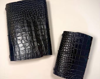 May include: Two dark navy blue journals with a glossy crocodile skin texture. Each notebook is secured with a thin black elastic band wrapped horizontally around the center, holding the covers closed in a rectangular shape.