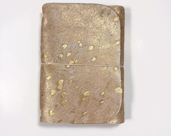 May include: A rectangular notebook with a textured, light brown hair-on-hide cover featuring irregular metallic gold splatters. The cover is secured by a thin, light-colored elastic band wrapped horizontally around the center of the notebook.