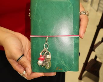 May include: A dark green leather journal with a textured finish, secured by a thin pink and white elastic band. A silver-toned metal charm hangs from the band, featuring a star shape, a white pearl bead, a small red accent, and a light green stone.