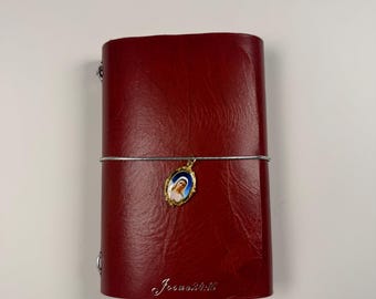 May include: A deep red leather journal with a textured surface, secured by a silver elastic band. An oval gold-toned charm featuring a depiction of the Virgin Mary hangs from the band. The text "Joshua 24:15" is embossed in silver at the bottom.