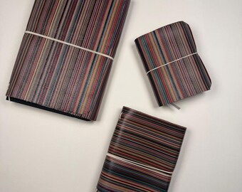 May include: Three rectangular notebooks featuring a multi-colored vertical striped pattern in shades of red, blue, yellow, and black. Each notebook is secured with a thin white elastic band wrapped horizontally around the center of the cover.