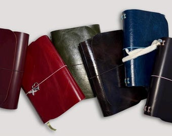 May include: A collection of six rectangular leather journals in various colors including burgundy, red, olive green, dark brown, navy blue, and black. Some journals feature elastic closures with metal heart and cross charms or white cord.