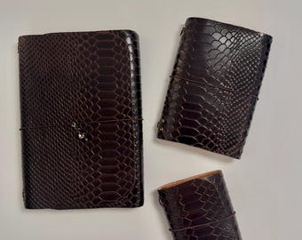 May include: Three dark brown journals with a textured snakeskin pattern. Each rectangular notebook is secured with a thin, dark elastic cord wrapped horizontally around the center. The journals vary in size, with one large, one medium, and one small.