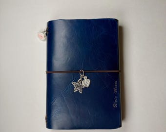 May include: A dark blue leather-like journal with a smooth, slightly crinkled texture. A thin brown elastic band wraps around the center, holding a silver star charm and a silver heart charm. The name Olivia Avans is embossed on the front cover.