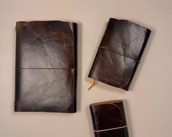 May include: Three rectangular journals with dark brown, textured leather covers. The largest journal is secured with a dark brown elastic band, the medium journal with a dark brown band, and the smallest journal with a thin pink elastic band.