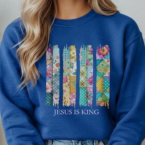 Puede incluir: Una sudadera de manga larga color azul real con un diseño gráfico de cuatro columnas verticales estilo pincelada, rellenas de coloridos patrones de patchwork florales y geométricos. El texto "JESUS IS KING" está impreso en blanco.