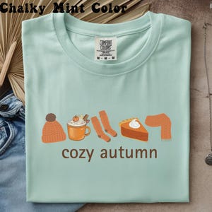 Puede incluir: Una camiseta de color verde menta tiza con un estampado gráfico de un gorro de punto naranja, una taza de café con leche de calabaza, dos calcetines naranjas, un trozo de tarta de calabaza y una bufanda naranja, con el texto marrón cozy autumn.