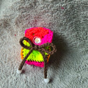 May include: A small, rectangular, crocheted pouch featuring vibrant neon pink, orange, and lime green yarn. The pouch is cinched with a dark brown crocheted drawstring tied in a bow, accented by a small white daisy button and white beads.