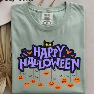 Puede incluir: Una camiseta de cuello redondo color verde salvia con un estampado gráfico que incluye el texto "HAPPY HALLOWEEN" en letras moradas. Sobre el texto hay un gato negro con ojos amarillos. Debajo, calabazas naranjas cuelgan de hilos.