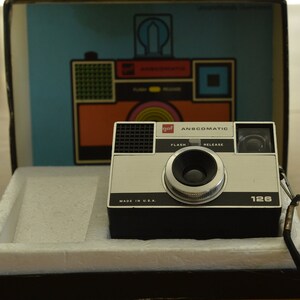 May include: A vintage black and silver 126 film camera with a red "gaf anscomatic" label. The camera has a flash and release button, a lens with a silver rim, and a black "made in u.s.a." label.