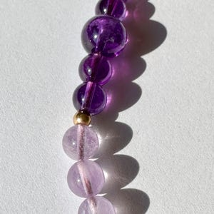 May include: A close-up of a purple and light pink amethyst gemstone beaded necklace. The beads are round and have a smooth, polished finish. The necklace is strung on a thin, gold-colored cord.