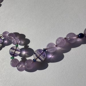May include: A purple and white striped gemstone necklace with smaller green and blue gemstones interspersed throughout.