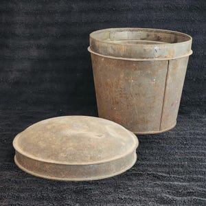 May include: An antique metal bucket and lid. The bucket is cylindrical with a slightly tapered shape and a rolled rim. The lid is round and domed, with a rolled edge. Both pieces have a weathered, rusty appearance.