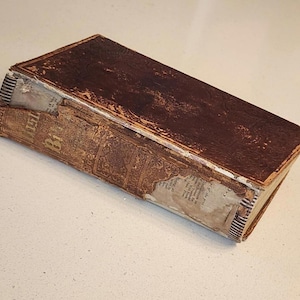 May include: An antique book with a worn, brown leather cover and aged, textured pages. The book's spine is partially visible, showing a striped pattern. The title "Bible" is visible on the side of the book.
