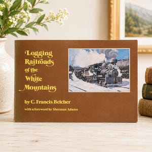 May include: A brown hardcover book titled Logging Railroads of the White Mountains by C. Francis Belcher with a foreword by Sherman Adams. The cover features a rectangular illustration of a steam locomotive on tracks in a snowy landscape.