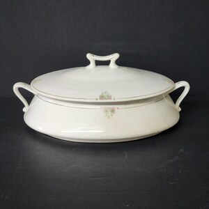 May include: A white porcelain serving dish with a lid. The dish has two handles and a handle on the lid. The dish is decorated with a gold trim and small floral accents in pink and green. The dish is on a black surface.