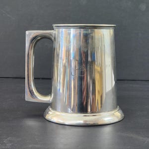 May include: A silver-colored tankard with a rectangular handle. The mug has a wide base and a slightly flared top. The initials "JB" are engraved on the front. The tankard is made of a shiny metal and reflects the surrounding light.