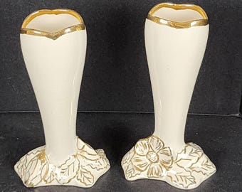 Könnte beinhalten: Zwei cremefarbene Blumenvasen mit goldenen Verzierungen und floralen Basen. Die Vasen haben eine ausgestellte Form und einen goldenen Rand. Die Basen weisen ein goldenes Blumendesign mit Blättern auf. Die Vasen sind wahrscheinlich aus Keramik oder Porzellan.