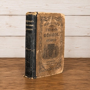 May include: An antique hardcover book titled Greenleaf's Common School Arithmetic with a black spine and a tan cover featuring black decorative borders, text, and a small illustration of figures standing near a large rectangular object.